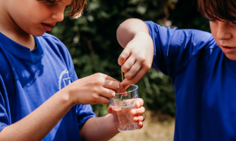 Two children work together on a science experiment outdoors, placing a dandelion into a clear plastic cup of water