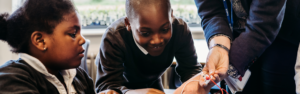 Two primary school children take part in a hands-on science activity while a teacher supports them, helping connect wires and components during a classroom investigation.