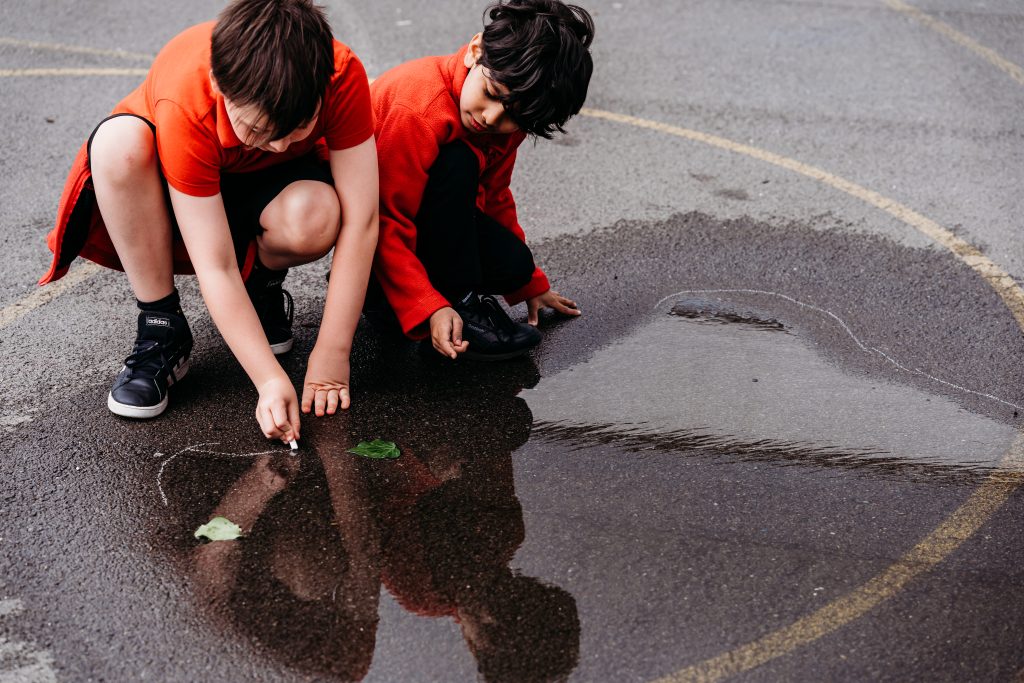 Two children drawing a chalk line around a puddle in the playground.