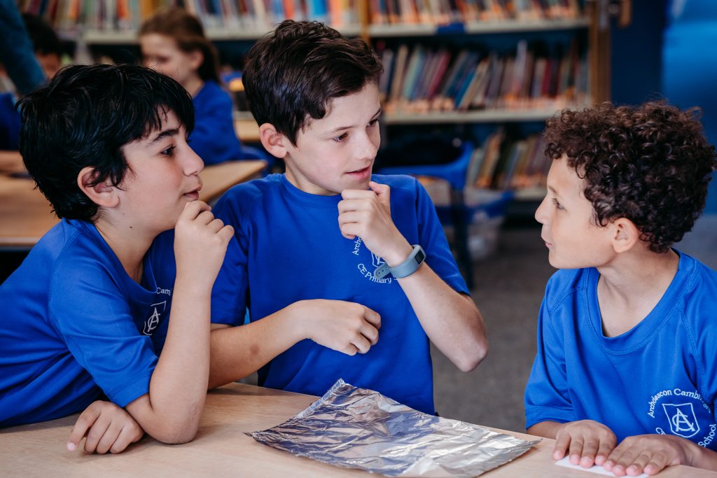 Three children sitting at a table in a classroom thinking and talking about the properties of foil.