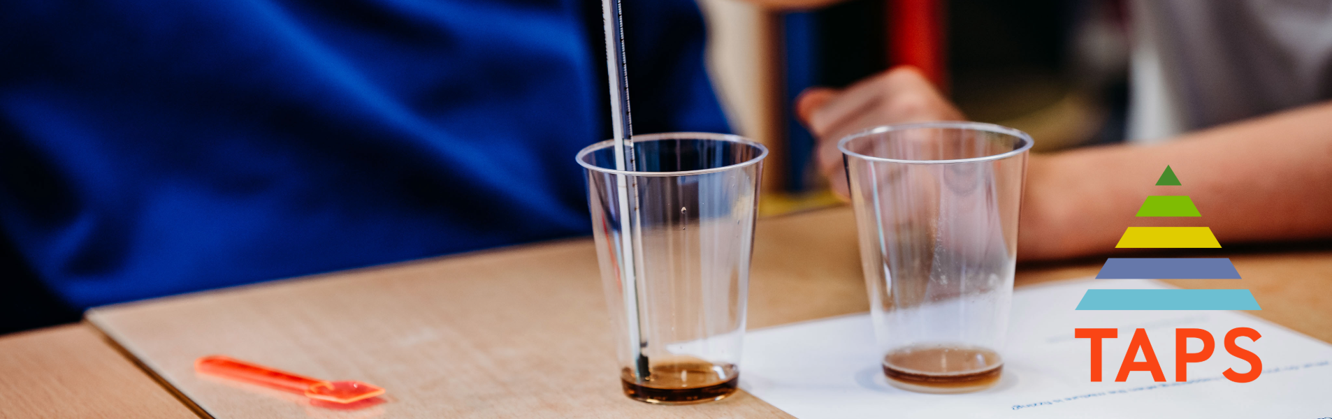children carrying out a science investigation using plastic cups, liquid and pipettes, with the TAPS logo in the right corner